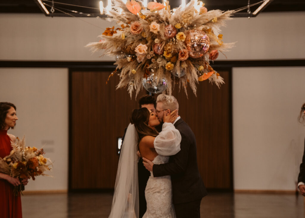 A bride and groom kissing above a terracotta, golden mustard, toffee, pampas disco ball flower installation at Meadville wedding venue The Harper.